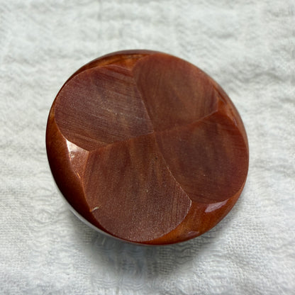 A round button with a carved geometric four-leaf pattern, made from reddish-brown casein with a glossy finish, resting on a textured white fabric background.